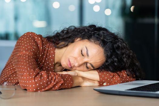 Beautiful tired hispanic woman sleeping on desk, close-up business woman with closed eyes napping inside office near laptop.