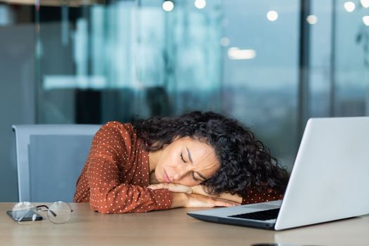 Beautiful tired hispanic woman sleeping on desk, close-up business woman with closed eyes napping inside office near laptop.