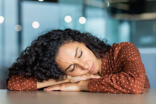 Beautiful tired hispanic woman sleeping on desk, close-up business woman with closed eyes napping inside office near laptop.