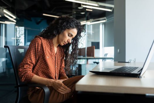 Latin American woman working inside office, business woman has severe leg pain, massaging muscle while sitting at table on chair, using laptop at work.