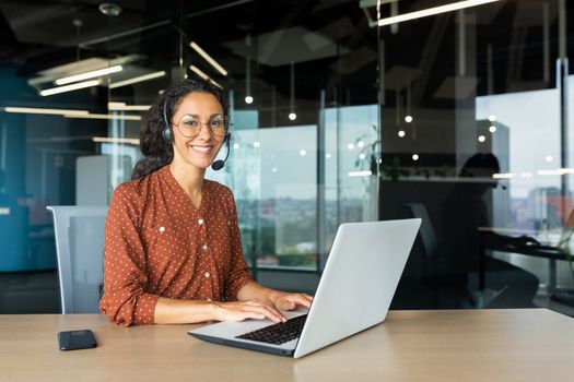 Portrait of Latin American business woman, office worker looking at camera and smiling, using headset and laptop for remote online communication, customer support tech call center worker.