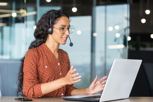 Video call online meeting with colleagues, Hispanic woman working inside modern office, businesswoman smiling and talking remotely using laptop and headset.