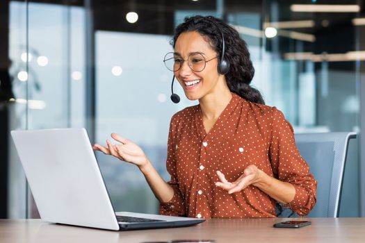 Video call online meeting with colleagues, Hispanic woman working inside modern office, businesswoman smiling and talking remotely using laptop and headset.