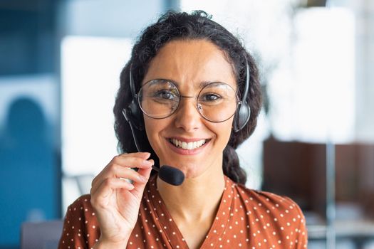 Close up portrait of latin american woman inside modern office with headset for video call, woman smiling and looking at camera, office worker customer support tech helpline.