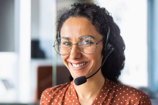 Close up portrait of latin american woman inside modern office with headset for video call, woman smiling and looking at camera, office worker customer support tech helpline.