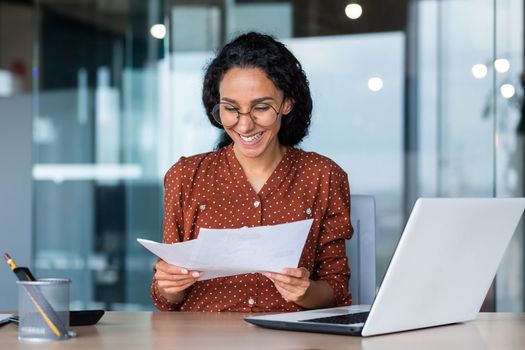 Young happy and successful businesswoman in glasses working with documents inside office, Hispanic woman with laptop looking at bills and contracts, financier with curly hair using laptop.