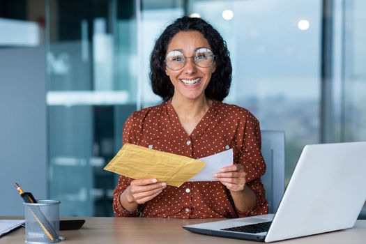Happy hispanic woman smiling and looking at camera, business woman received envelope with good news notification letter, office worker with documents and laptop working in office at work.