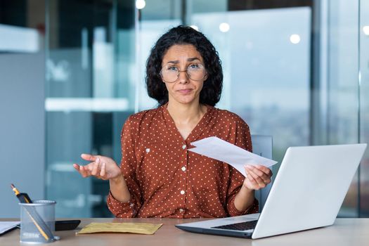 Portrait of disappointed business woman, latin american received notification letter from bank with bad news, office worker looking sad at camera, working at work using laptop.