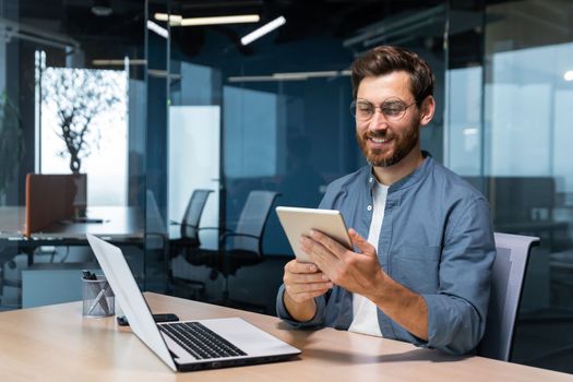 Successful businessman in a shirt uses a tablet computer, a man sits at a desk smiling and happy, a programmer tests a new application.