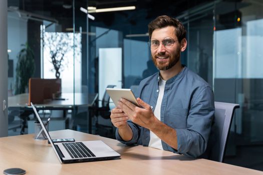 Portrait of a businessman in a modern office, a man uses a tablet computer, sits at a desk, smiles and looks at the camera in a casual shirt and glasses.