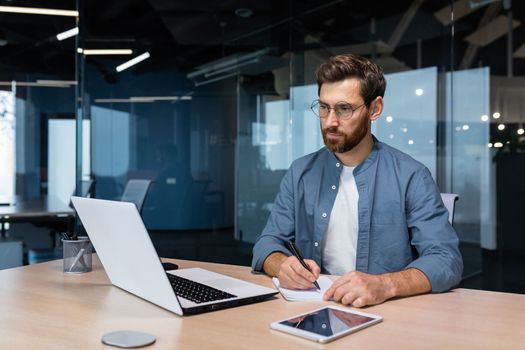 Serious businessman is writing down information studying online while sitting inside the office.