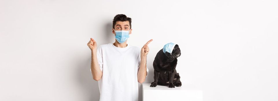 Covid-19, animals and quarantine concept. Young man and black dog wearing medical masks, pug looking at upper left corner and owner showing promo offer, rejoicing.