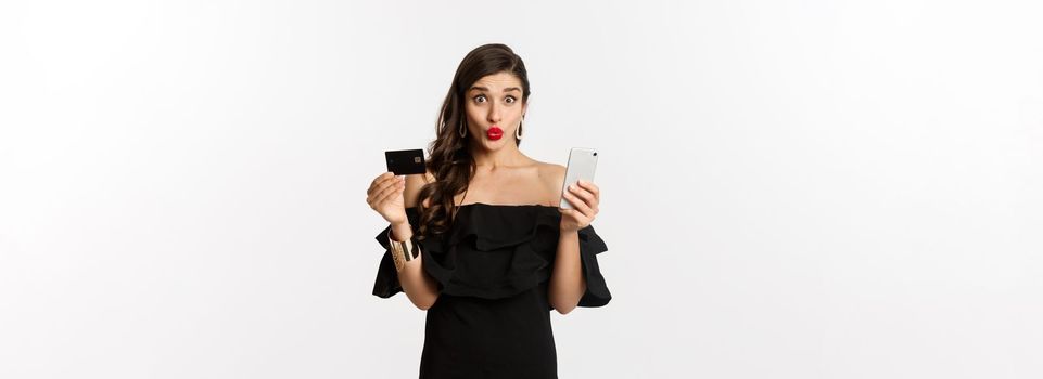 Online shopping concept. Fashionable woman in black dress, holding credit card with smartphone, looking excited, standing over white background.