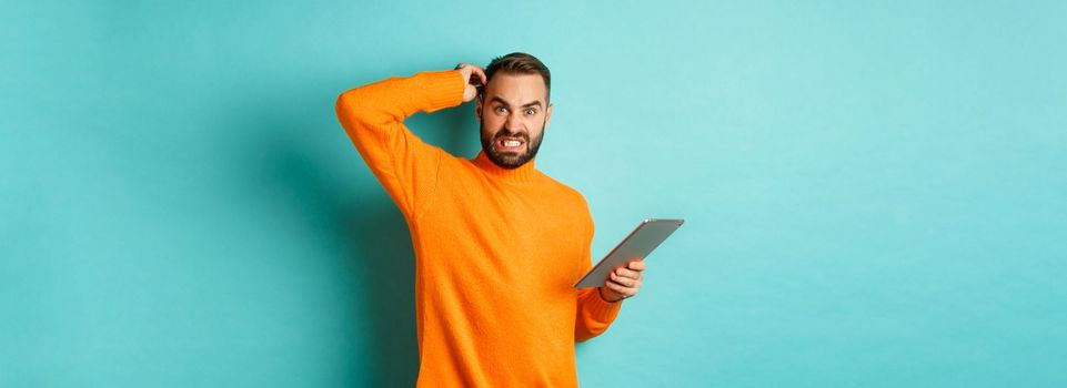 Confused man scratching head and looking worried, working on digital tablet, standing against turquoise background.