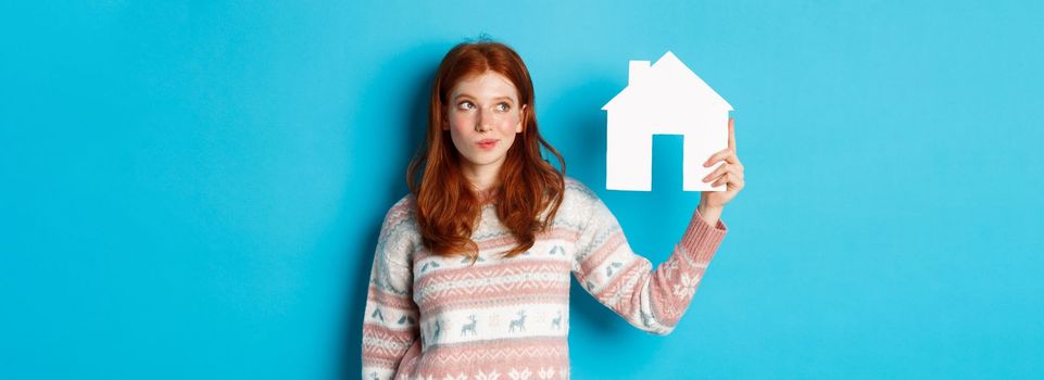 Real estate concept. Image of thoughtful redhead girl showing paper house model and thinking, searching for home or flat, standing against blue background.