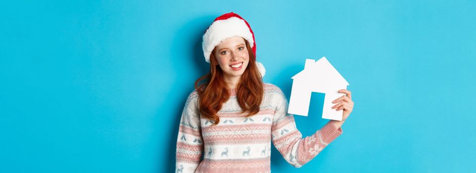 Holiday promos and real estate concept. Cheerful redhead woman in santa hat holding paper house in hand and smiling, standing in sweater against blue background.