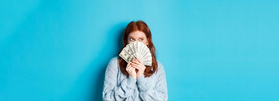 Thoughtful cute girl with red hair dreaming about shopping, holding dollars and looking at upper left corner logo, standing over blue background.