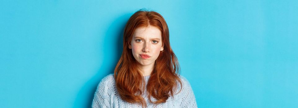 Disappointed teenage girl with red hair, frowning and smirking displeased, looking judgemental, standing against blue background.