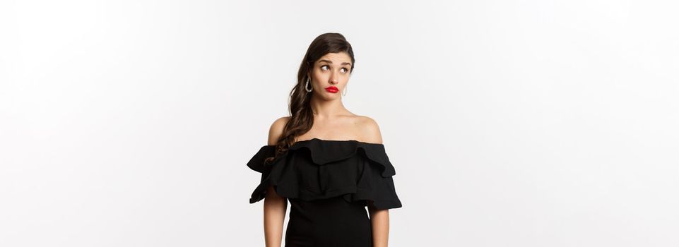 Fashion and beauty. Confused attractive woman in black dress, looking away with puzzled and shocked face, standing over white background.