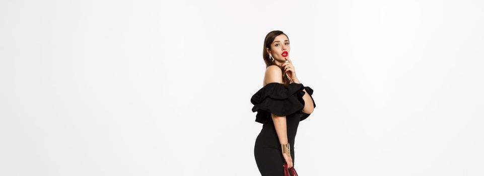 Beauty and fashion concept. Full length of sensual young woman in black elegant dress, high heels and purse, touching lip flirty and gazing at camera, white background.