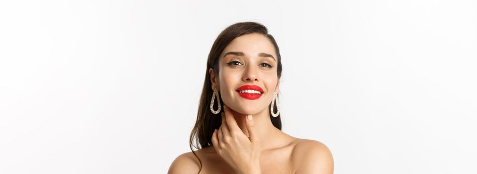 Fashion and beauty concept. Close-up of gorgeous brunette woman with red lips, touching face and smiling self-assured, standing over white background.