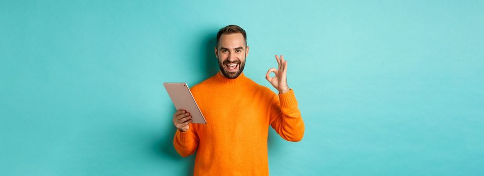 Satisfied adult man smiling, using digital tablet and showing okay sign, approve and agree, standing against turquoise background.