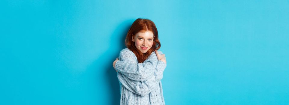 Happy cute redhead girl hugging herself, wearing comfortable and warm sweater, smiling at camera, standing over blue background.