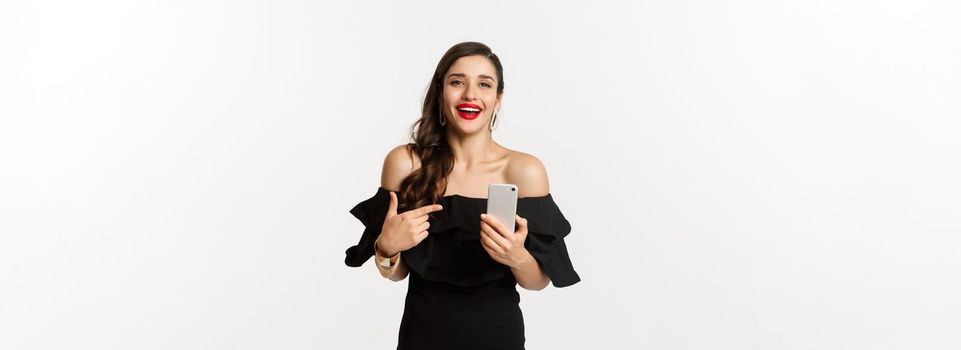 Online shopping concept. Satisfied pretty woman in black dress, smiling pleased and pointing at mobile phone, standing over white background.