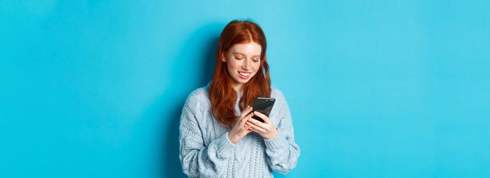 Young teenage redhead girl reading message on smartphone and smiling, using mobile phone and standing over blue background.