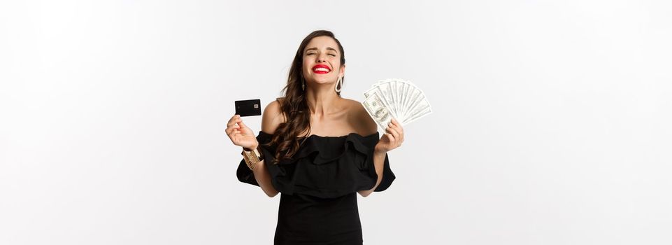 Beauty and shopping concept. Satisfied young woman in stylish dress, looking pleased, holding credit card and money, standing over white background.