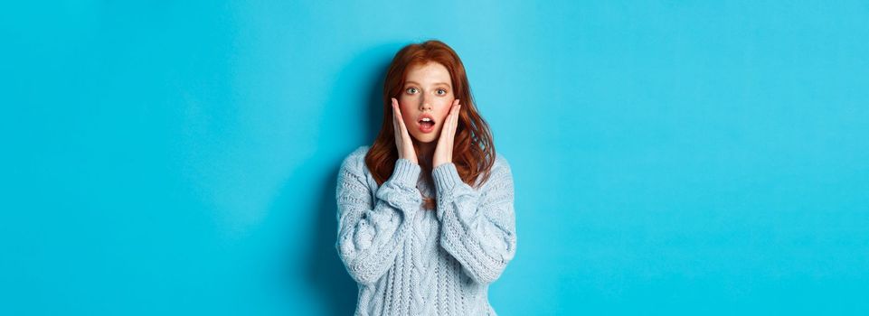 Image of young beautiful redhead girl, looking surprised, staring with complete disbelief at camera, standing in sweater against blue background.