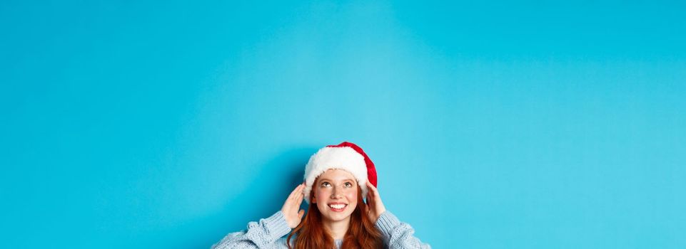 Winter holidays and Christmas eve concept. Head of cute redhead girl in santa hat, appear from bottom and looking up at copy space, staring logo, standing over blue background.