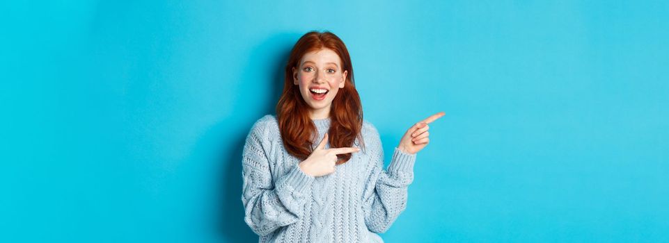 Amazed teenage girl with red hair and freckles, pointing fingers left at logo and smiling, showing advertisement, standing over blue background.