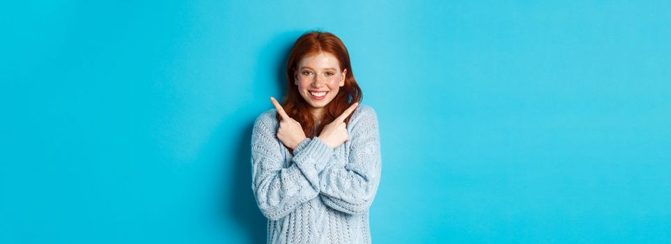 Cheerful redhead girl showing two choices, pointing sideways and smiling over blue background. Copy space