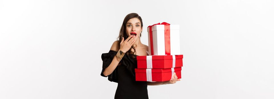 Celebration and christmas holidays concept. Woman holding xmas gifts and looking surprised, receive presents, standing over white background.