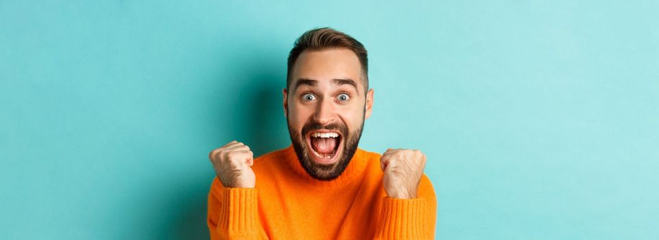 Image of handsome relieved man feeling satisfaction, rejoicing of winning or achievement, making fist pump and saying yes, standing over turquoise background.