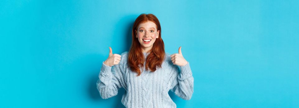 Cheerful teen girl with red hair, showing thumbs up in approval, like and praise gesture, standing over blue background.