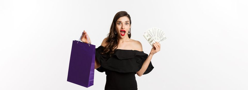 Happy woman buyer holding shopping bag and money, standing in black dress over white background. Copy space