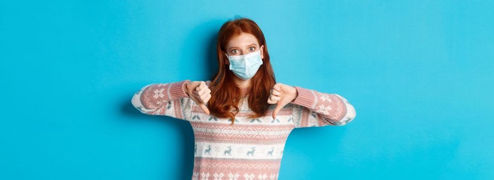 Winter, covid-19 and pandemic concept. Displeased redhead woman in medical mask, showing thumbs-down in dislike, disagree with you, standing over blue background.