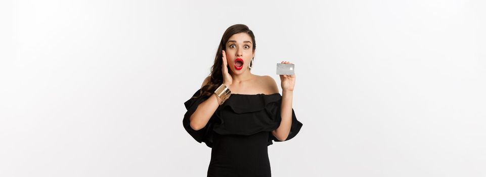 Fashion and shopping concept. Excited elegant woman with red lips, wearing black dress, showing credit card and looking amazed, standing over white background.