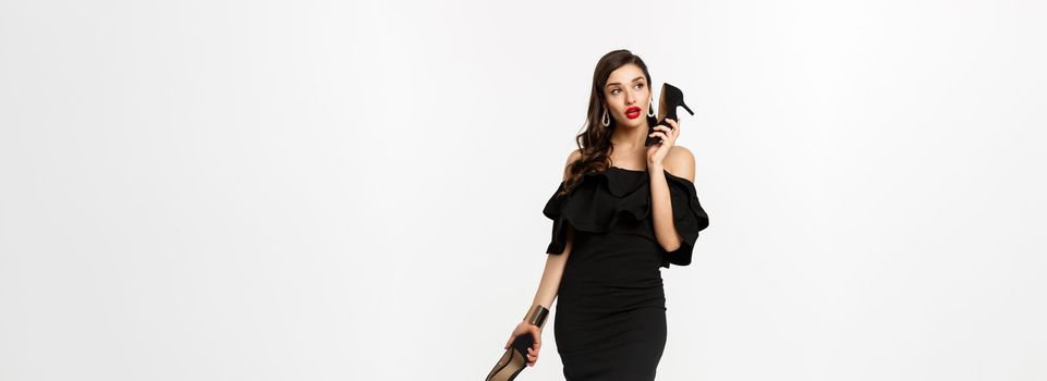 Beauty and fashion concept. Full length of attractive young woman using high heels like mobile phone, standing in black dress against white background.