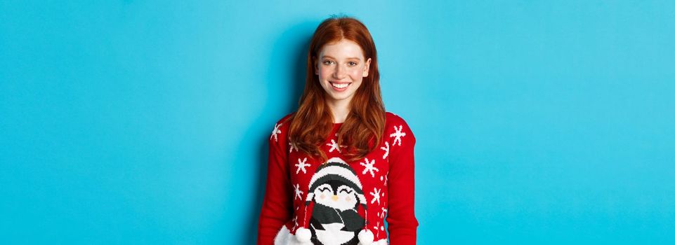 Winter holidays and Christmas Eve concept. Cute smiling teenage girl with red hair, wearing funny xmas sweater, standing against blue background.