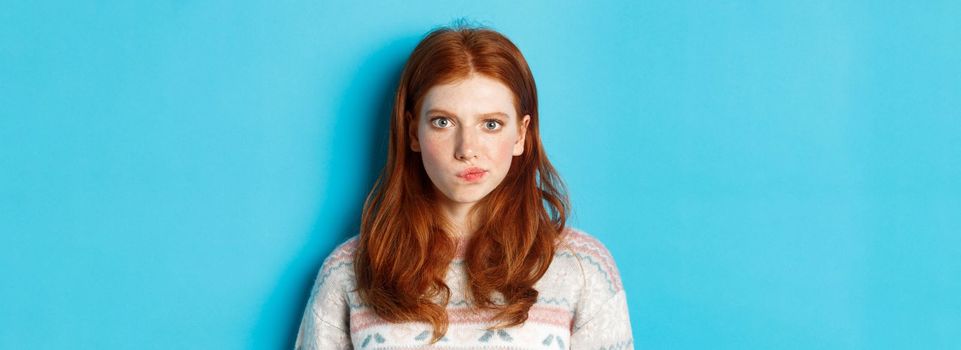 Close-up of serious-looking girl thinking, staring at camera troubled and grimacing, making hard choice, standing over blue background.