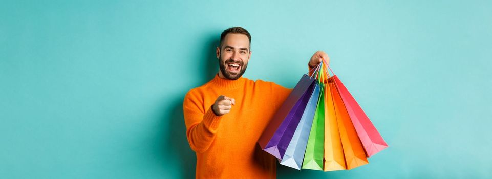 Happy adult man pointing finger at camera, holding shopping bags and smiling, standing over turquoise background.