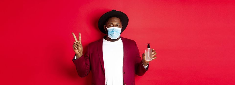 Covid-19, quarantine and holidays concept. Young african american man in medical mask showing hand sanitizer and peace sign, recommending to use antiseptic.