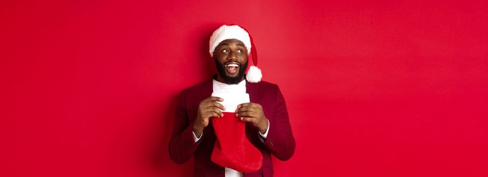 Cheerful Black man looking upper left corner and smiling, holding Christmas sock with gifts, standing over red background.