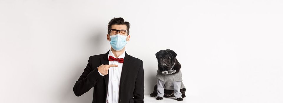 Coronavirus, pets and celebration concept. Disappointed young man in face mask and suit, pointing finger at cute black pug dog wearing party costume, standing over white background.