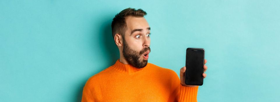 Close-up of young bearded man showing phone screen and looking amazed, wearing orange sweater, standing against studio background.