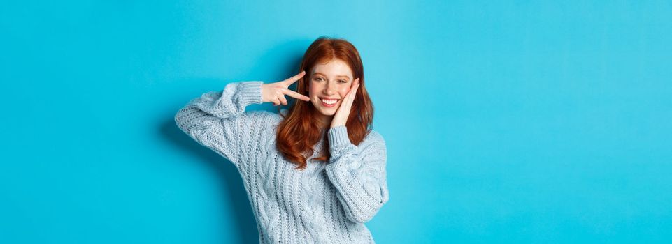 Cheerful redhead female model sending good vibes, smiling and showing peace sign, standing over blue background.