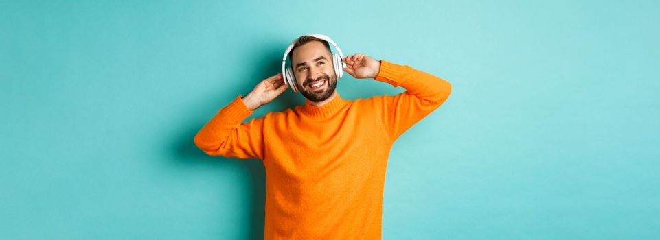 Handsome bearded man listening to music in headphones, smiling pleased, standing over turquoise background.